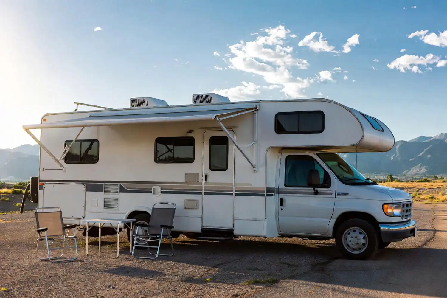 White Class C motorhome parked at a campground site with mountain views, an awning extended, and two folding chairs outside