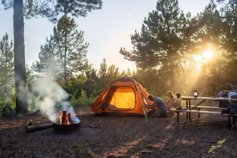 Family setting up a dome tent at a forested campground site at golden hour with a small campfire and picnic table