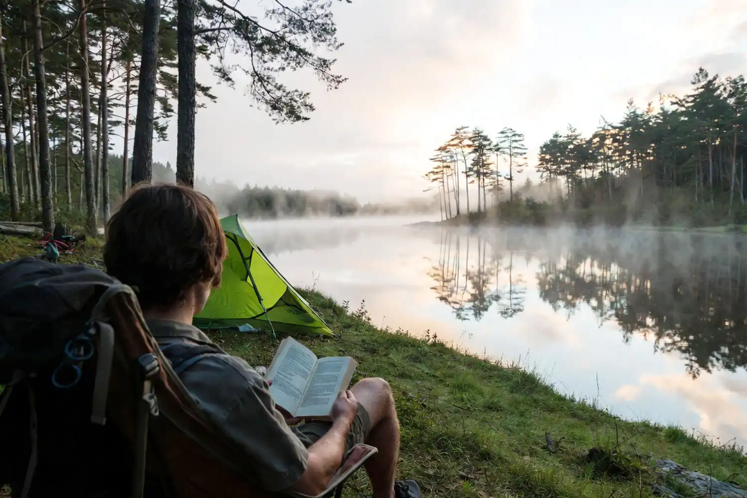 Lone camper sitting in a folding chair reading a book beside a calm lake with a small tent